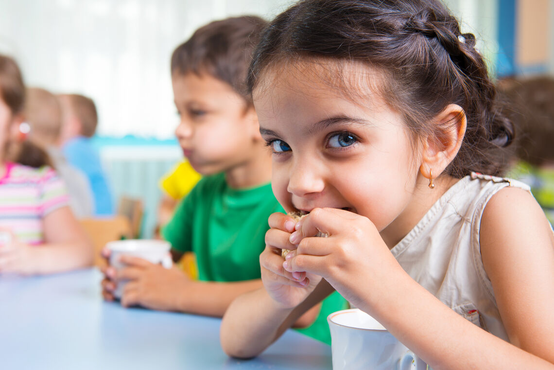 young girl biting into lunch in school cafeteria and looking at camera